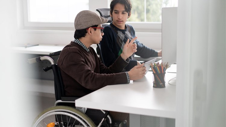 A guy in a wheelchair studying and discussing with another guy in front of a computer